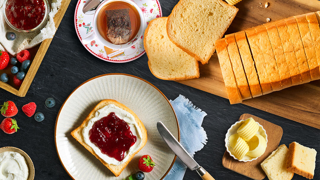 Frühstücksszene mit vorgeschnittener Brioche auf einem Brettchen, einer Scheibe bestrichen mit weißer Creme und roter Marmelade, umgeben von Butterstücken, frischen Beeren, Marmeladenglas und einer Tasse Tee.