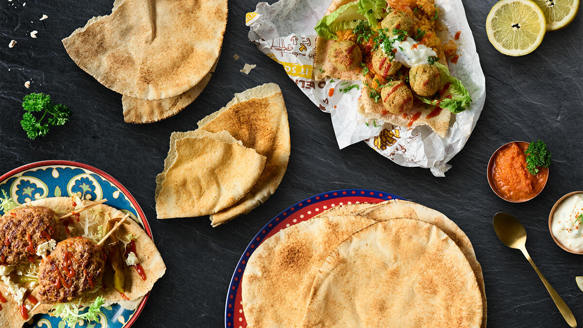Mehrere dünne, runde arabische Fladenbrote auf bunten Tellern, dazu gefüllte Wraps mit Falafel, Salat und Sauce. Im Bild außerdem Zitronenscheiben, rote Paprika-Dip und Kräuter-Dip in kleinen Schalen.