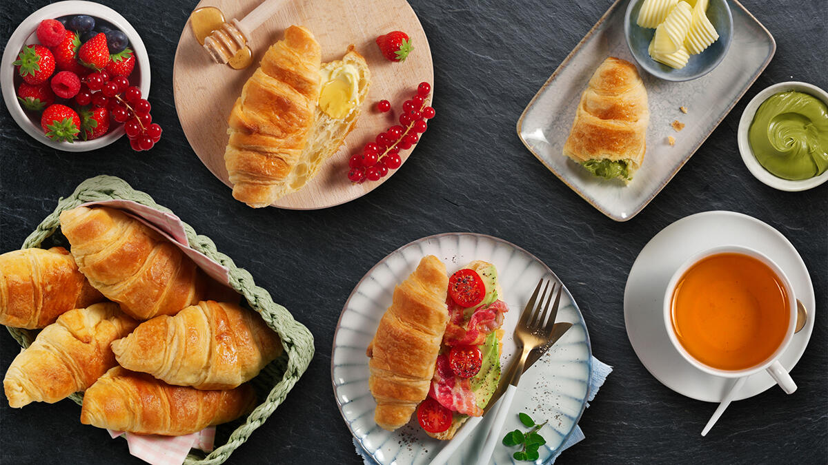 Das Bild zeigt mehrere goldbraune Croissants in einer Frühstücksszene. Links liegt ein Korb voller Croissants. Oben ein Croissant auf einem Holzbrett, aufgeschnitten und mit Butter und Honig bestrichen, daneben rote Beeren. Rechts ein Teller mit einem Croissant-Stück und grüner Creme sowie Butterstückchen. Unten ein Teller mit einem herzhaft belegten Croissant mit Avocado, Tomaten und Schinken. Außerdem sind frische Beeren, eine Schale mit grüner Creme und eine Tasse Tee zu sehen.