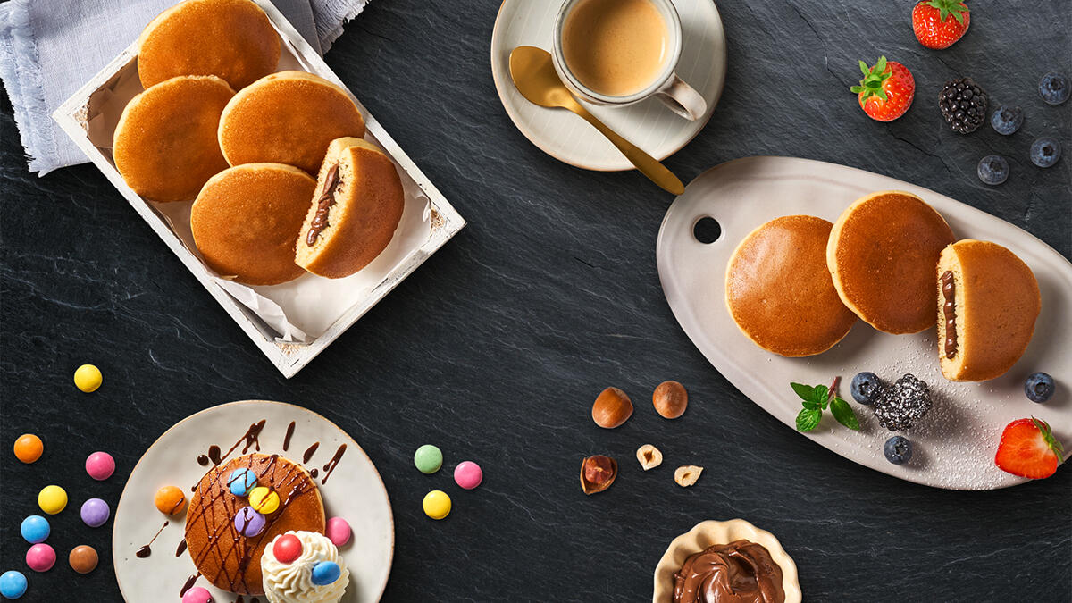 Das Bild zeigt mehrere goldbraune Pancakes mit Schokoladenfüllung. Links liegt eine weiße Schachtel mit Pancakes, einer ist halbiert und zeigt die Füllung. Rechts auf einem Teller liegen drei weitere Pancakes, einer aufgeschnitten. Rundherum sind frische Beeren, bunte Schokolinsen, Haselnüsse, eine Schale mit Schokoladencreme, eine Tasse Kaffee und ein dekorierter Pancake mit Sahne und Schokosoße.