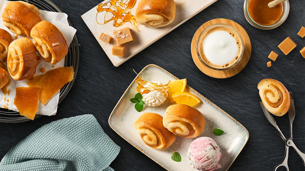 Mehrere Mini Schnecken mit Toffe-Geschmack auf einem Teller mit einer Kugel Eis, Orangenspalten und Sahnetupfern. Im Bild außerdem ein Gitter mit weiteren Schnecken, Toffee-Stückchen, Karamellsoße im Glas und eine Tasse Milchkaffee mit Milchschaum.