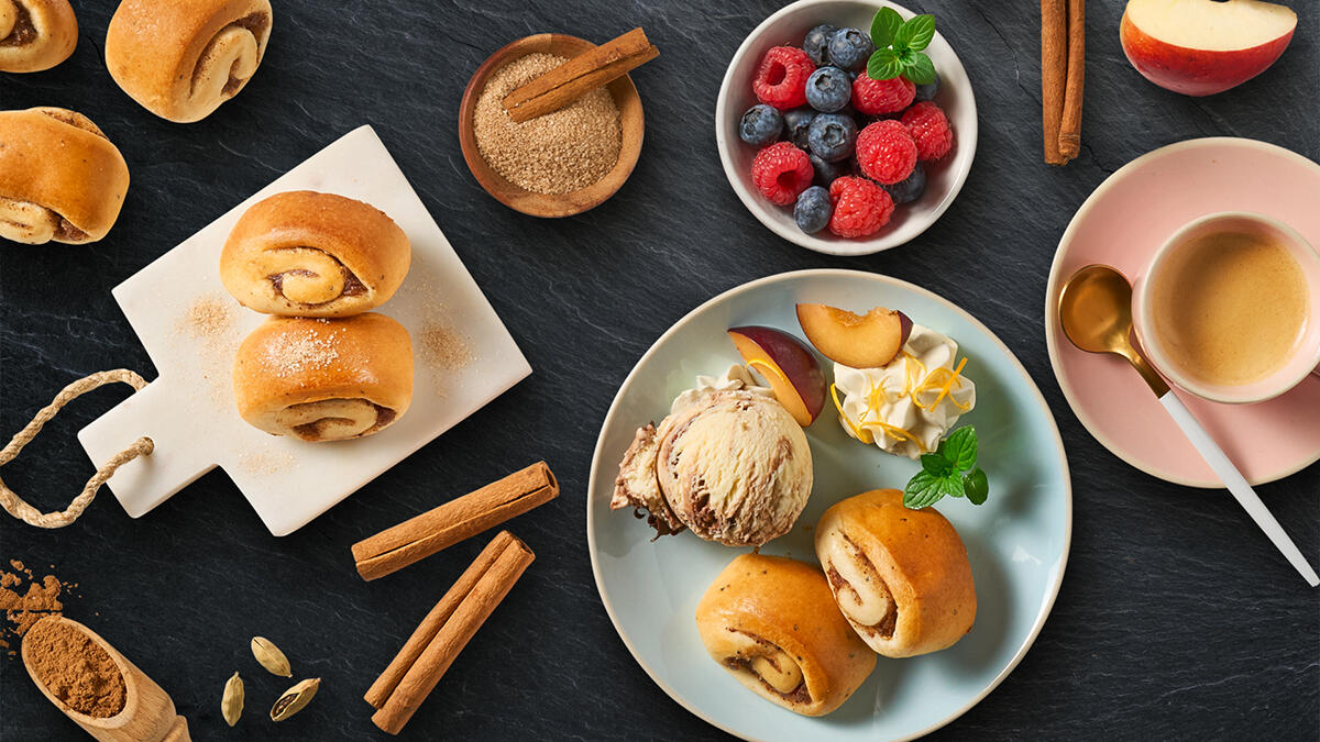 Mehrere kleine Schnecken auf einem Schneidebrett und einem Teller, begleitet von einer Kugel Eis, frischen Pfirsichstücken und Sahnetupfern. Im Bild außerdem eine Schale mit Beeren, eine Tasse Espresso, Zimtstangen, gemahlener Zimt und Kardamom.