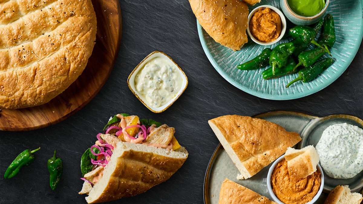 Mehrere angerichtete Teller mit frisch gebackenem Fladenbrot und verschiedenen Dips auf dunkler Steinoberfläche. Links ein ganzes rundes Fladenbrot auf Holzbrett, daneben ein Sandwich mit gegrilltem Fleisch, roten Zwiebeln und Gemüse. Rechts oben ein Teller mit Fladenbrotstücken, grünen Pimientos und zwei Schälchen mit grünem und rotem Dip. Unten rechts ein weiterer Teller mit Fladenbrotstücken, rotem Dip und einer Schale mit Kräuter-Dip. Die Szene wirkt mediterran und appetitlich.