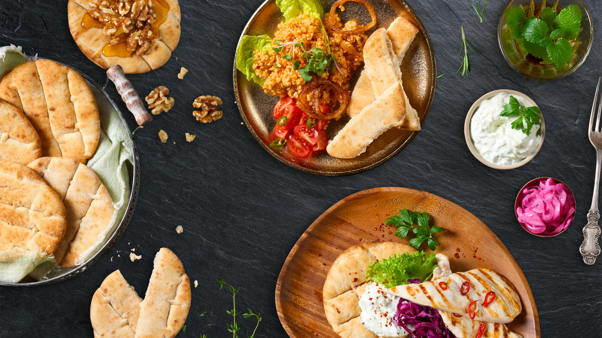 Verschiedene mediterrane Gerichte auf dunkler Steinplatte: Mini-Pitas, angerichtet mit frischen Beilagen wie Tomaten, Bulgur-Salat, gegrilltem Käse, Kräutern und Dips wie Tzatziki. Dazu eingelegte rote Zwiebeln und Walnüsse als Garnitur