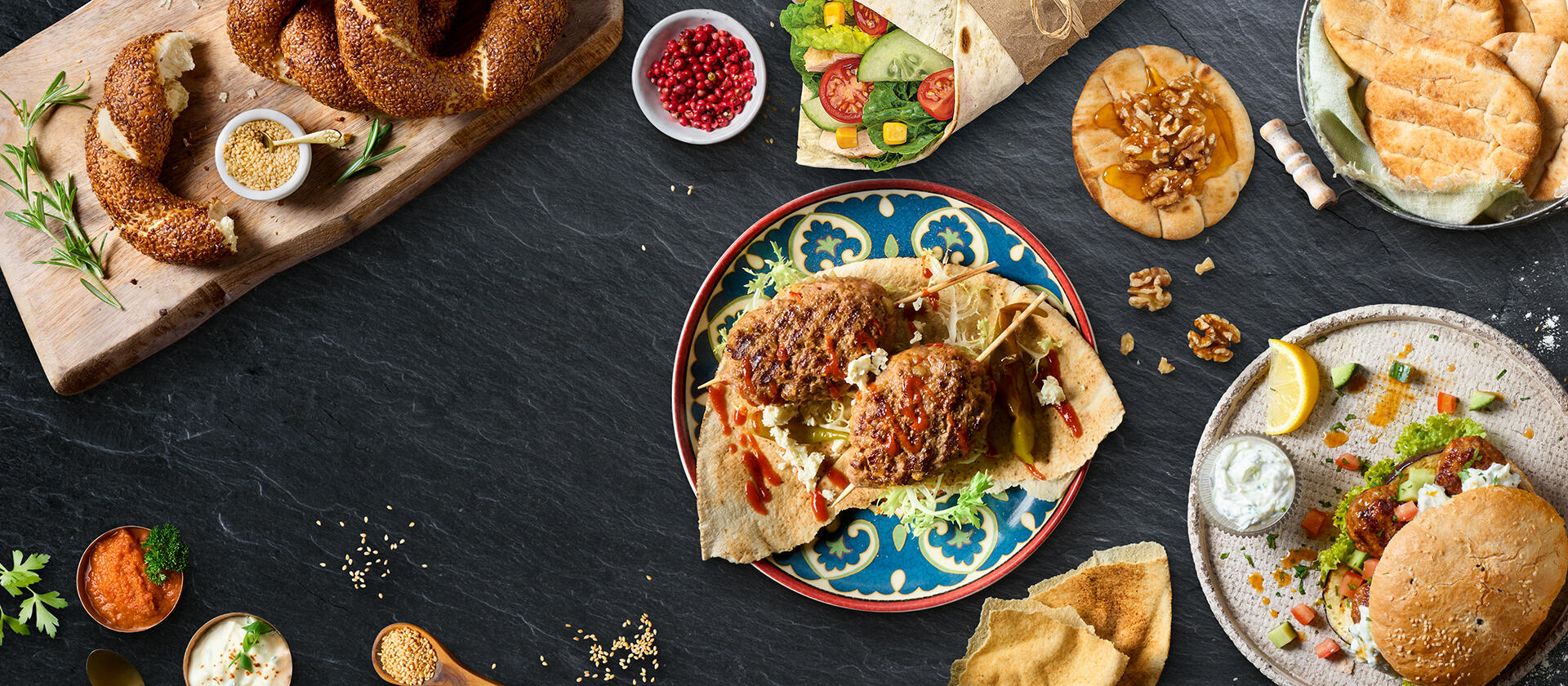 Orientalische Speisen auf dunkler Steinplatte: Fladenbrot mit Fleischspießen, Sesamringe, Wrap mit Gemüse, Mini Pitas und Mini Fladenbrote mit Falafel, dazu Dips, Gewürze und Kräuter.